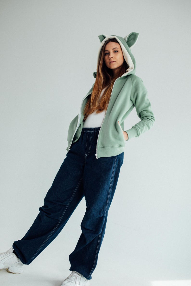 May include: A light green hooded sweatshirt with white fur lining the hood and ears. The sweatshirt has a zipper closure and is worn with dark blue jeans.