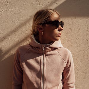 May include: A woman wearing a light pink hooded sweatshirt with a zipper. She is wearing sunglasses and looking to the right.