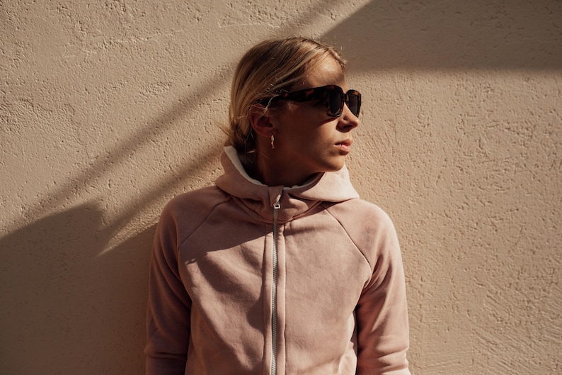 May include: A woman wearing a light pink hooded sweatshirt with a zipper. She is wearing sunglasses and looking to the right.