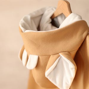 K&ouml;nnte beinhalten: Ein beiger Kapuzenpullover mit wei&szlig;en Ohren an der Kapuze. Der Pullover h&auml;ngt an einem Holzb&uuml;gel.
