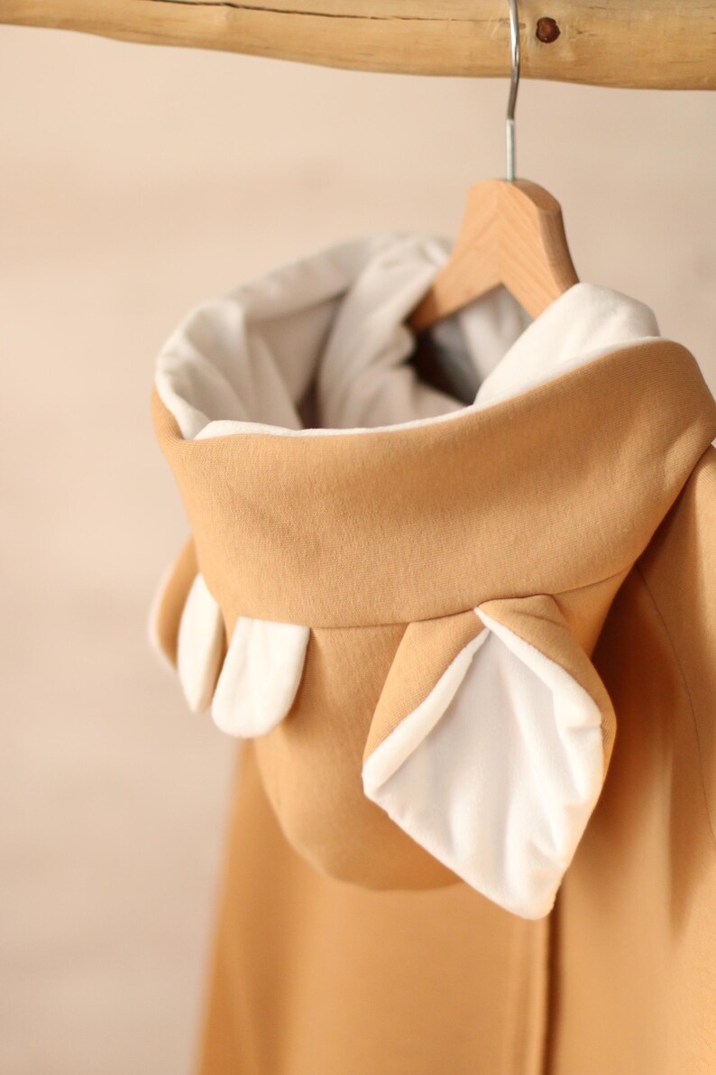 K&ouml;nnte beinhalten: Ein beiger Kapuzenpullover mit wei&szlig;en Ohren an der Kapuze. Der Pullover h&auml;ngt an einem Holzb&uuml;gel.