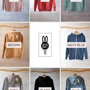 May include: Nine different color hooded sweatshirts with bunny ears on the hood. The colors are pink, red, green, brown, navy blue, grey, black, and blue. The sweatshirts are hanging on wooden hangers.