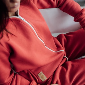 K&ouml;nnte beinhalten: Ein roter Kapuzenpullover mit Rei&szlig;verschluss und eine passende Jogginghose mit einem kleinen wei&szlig;en Logo auf der Tasche. Das Logo zeigt ein Kaninchen mit einem Herzen.