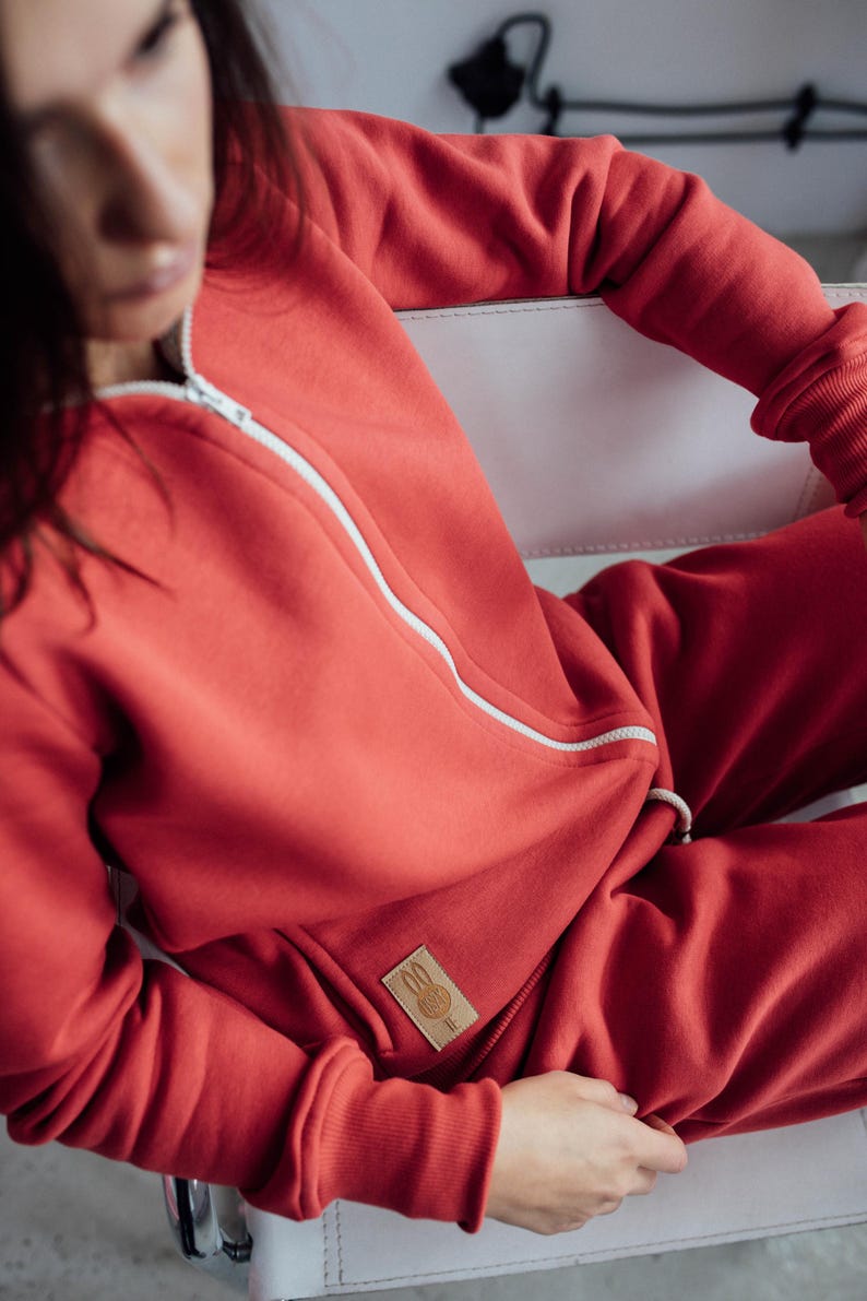K&ouml;nnte beinhalten: Ein roter Kapuzenpullover mit Rei&szlig;verschluss und eine passende Jogginghose mit einem kleinen wei&szlig;en Logo auf der Tasche. Das Logo zeigt ein Kaninchen mit einem Herzen.