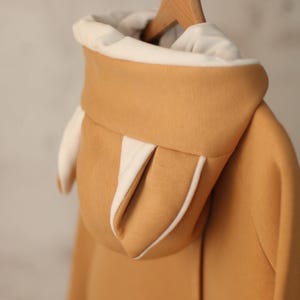 May include: A tan hoodie with a white lining on the hood. The hood has two small, white, pointed ears.