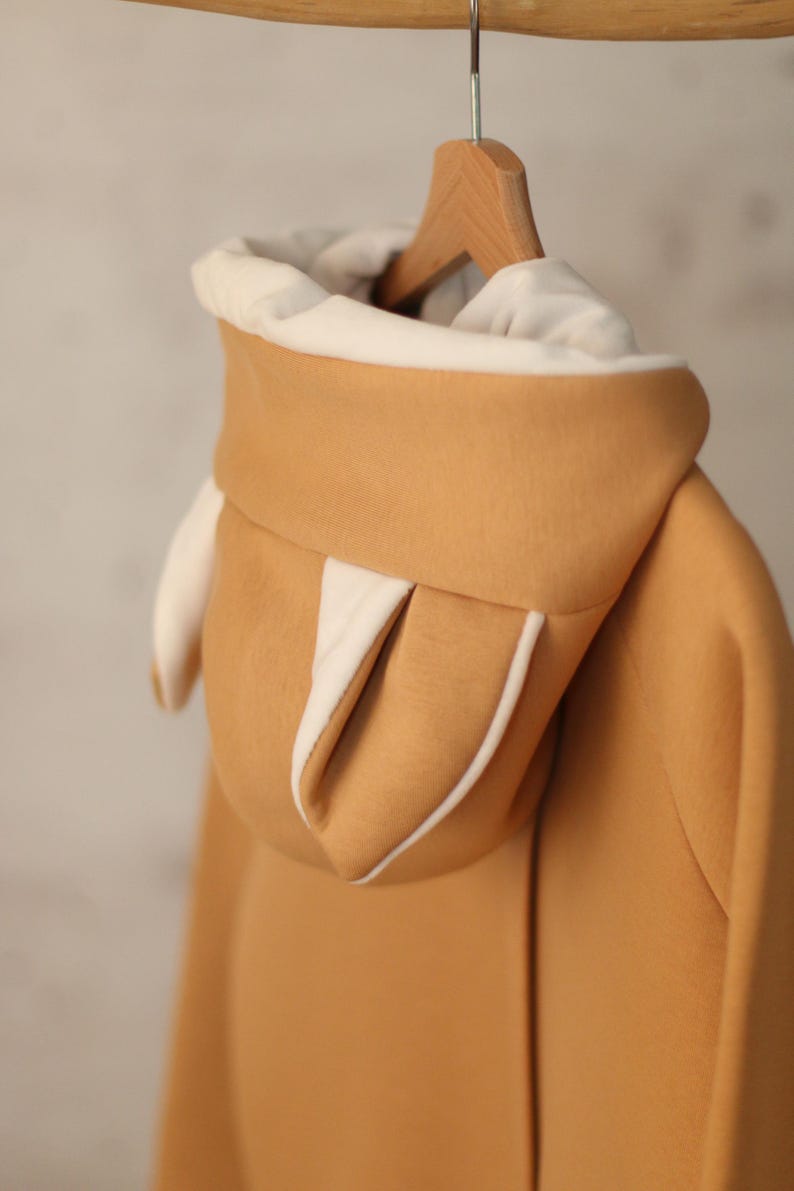 May include: A tan hoodie with a white lining on the hood. The hood has two small, white, pointed ears.