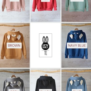 K&ouml;nnte beinhalten: Neun Kapuzenpullover in verschiedenen Farben mit Hasenohren an der Kapuze. Die Farben sind rosa, rot, gr&uuml;n, braun, marineblau, grau, schwarz und blau. Der Text "USZY TE" befindet sich in der Mitte des Bildes.