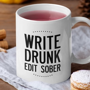 May include: White ceramic mug with the text "WRITE DRUNK EDIT SOBER" in black. The mug is filled with a pink liquid. The mug is on a white cloth, with cinnamon sticks and a pastry in the background.