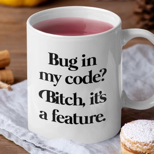 May include: White ceramic mug with the text "Bug in my code? Bitch, it's a feature" in black. The mug is filled with a pink liquid. The mug is on a white cloth with a pastry and cinnamon sticks in the background.