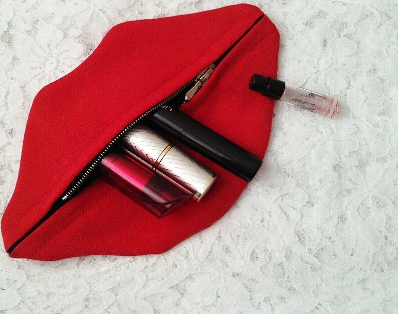 May include: A red felt zippered pouch shaped like a pair of lips, open to reveal a lipstick, a lip gloss, a mascara, and a small bottle of perfume.