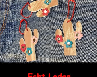 May include: Three light brown leather cactus-shaped ornaments with red, blue, and pink flower-shaped accents attached with metal rivets. Each cactus features a red ball chain loop for hanging. The text Echt Leder is printed in red at the bottom.