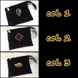 May include: Three black velvet pouches with different colored embellishments. The first pouch has a green teardrop-shaped embellishment. The second pouch has a purple square-shaped embellishment. The third pouch has a gold teardrop-shaped embellishment.