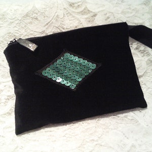 May include: A black velvet pouch with a green sequin diamond shape in the center. The pouch has a zipper closure and a black strap.