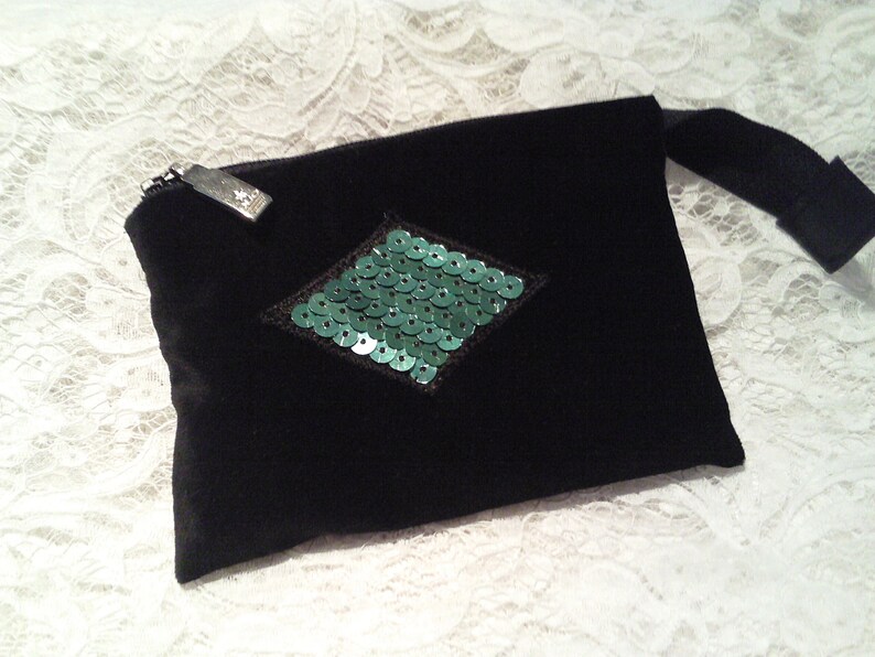 May include: A black velvet pouch with a green sequin diamond shape in the center. The pouch has a zipper closure and a black strap.