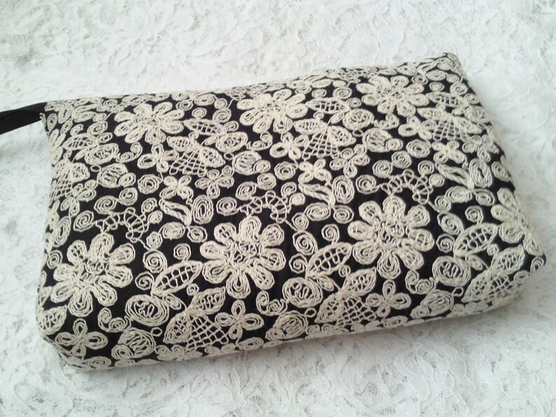 Op de afbeelding: Zwarte clutch met een wit kantpatroon. De tas heeft een ritssluiting en een klein handvat.