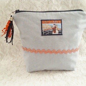 Op de afbeelding: Een lichtblauwe ritszak met een rode en witte rand. De tas heeft een klein, zilveren belletje met een zwart en oranje kwastje. De tas heeft een vintage-stijl patch met de tekst "Visit America" met een afbeelding van een vrouw op een schip.