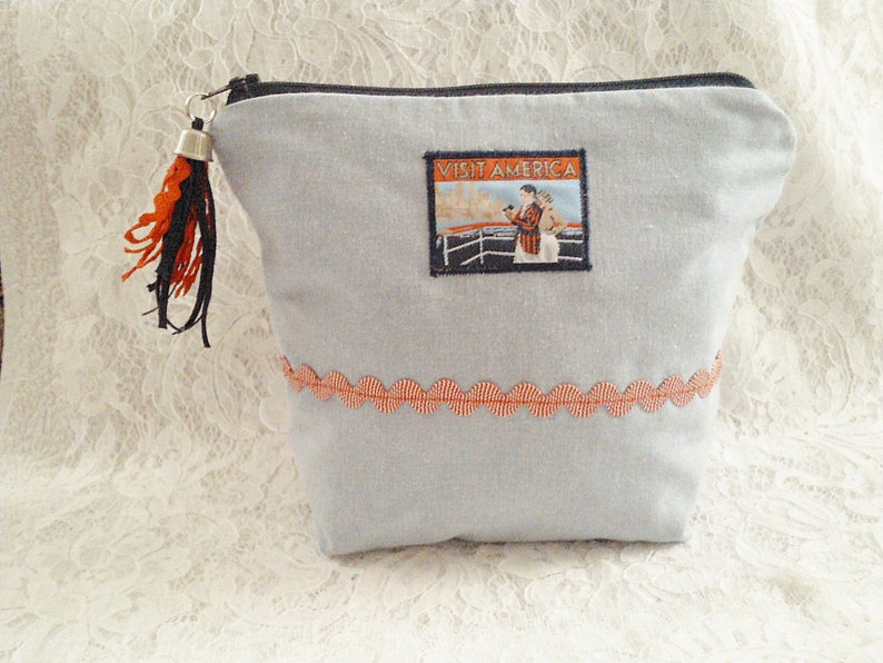 Op de afbeelding: Een lichtblauwe ritszak met een rode en witte rand. De tas heeft een klein, zilveren belletje met een zwart en oranje kwastje. De tas heeft een vintage-stijl patch met de tekst "Visit America" met een afbeelding van een vrouw op een schip.