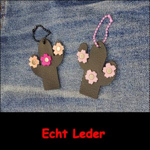 May include: Two dark brown cactus-shaped leather keychains with flower-shaped embellishments. One features a black ball chain and pink and peach flowers, while the other has a light purple ball chain and light pink flowers. Text reads Echt Leder.