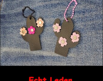 May include: Two dark brown cactus-shaped leather keychains with flower-shaped embellishments. One features a black ball chain and pink and peach flowers, while the other has a light purple ball chain and light pink flowers. Text reads Echt Leder.