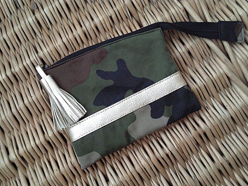 May include: A small green, brown, and black camouflage fabric pouch with a gold leather trim and a gold tassel. The pouch has a black zipper and a black strap.