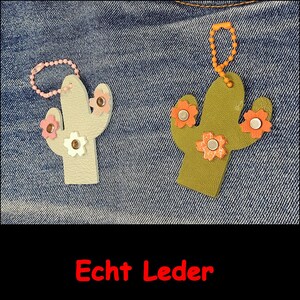 May include: Two cactus-shaped keychains made of textured material. The left one is light gray with pink and white flower accents and a light pink ball chain. The right one is olive green with orange flower accents and an orange ball chain.
