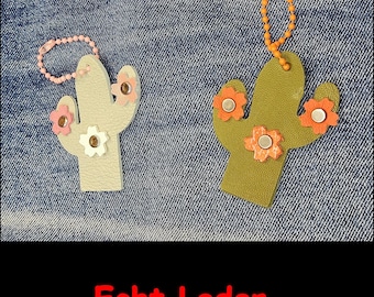 May include: Two cactus-shaped keychains made of textured material. The left one is light gray with pink and white flower accents and a light pink ball chain. The right one is olive green with orange flower accents and an orange ball chain.