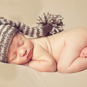 May include: A newborn baby wearing a brown and white striped knitted hat with a long tail and a pom-pom on top. The baby is sleeping on a beige surface.