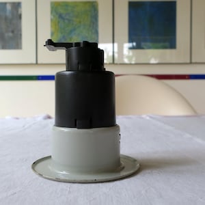 May include: A black and white industrial-style object with a circular base. The top portion is black, cylindrical, and has a small arm extending from it. The base is a light gray color. The object sits on a white surface.