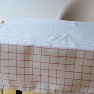Könnte beinhalten: Eine weiße Tischdecke mit rotem Karomuster bedeckt einen Tisch. Der Stoff drapiert über die Kanten, und das Muster ist gleichmäßig verteilt. Der Tisch steht vor einem neutralen Hintergrund, was auf einen Ess- oder Küchenbereich hindeutet.