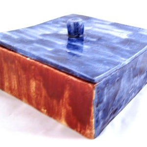 Può includere: Una scatola in ceramica con un coperchio. La scatola è rossa con un coperchio blu. Il coperchio ha un piccolo pomo in cima.