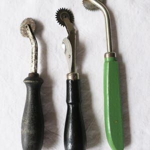 Puede incluir: Tres herramientas de costura vintage con ruedas metálicas y mangos de madera. Los mangos son negros y verdes. Las herramientas están dispuestas sobre una superficie blanca. Las ruedas tienen un borde dentado.