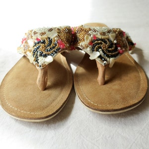 May include: A pair of brown leather sandals with a beaded embellishment on the toe strap. The embellishment is made of white, gold, and black beads, with a few red beads scattered throughout.