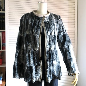 Puede incluir: Chaqueta de piel sintética gris y negra con cuello redondo y mangas largas. La chaqueta tiene un ribete de color claro y está abierta por delante. La chaqueta se exhibe en un maniquí.