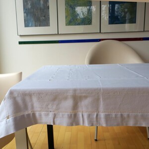Puede incluir: Un mantel rectangular blanco con detalles bordados cubre una mesa. La mesa está en una habitación con obras de arte en la pared y dos sillas de color crema. El mantel tiene un borde decorativo.