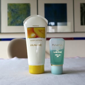 Könnte beinhalten: Zwei Tuben Lotion, eine weiße mit gelbem Deckel und den Worten "Mango Body Lotion Rabe" auf der Vorderseite und die andere blaue mit goldenem Deckel und den Worten "Plantess Gel Après Soleil after sun gel" auf der Vorderseite.