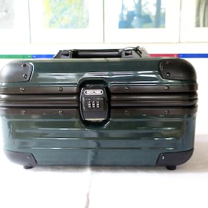 Può includere: Una valigia da cabina Rimowa verde scuro con manico e protezioni angolari neri. La valigia ha una serratura a combinazione ed è progettata per i viaggi.