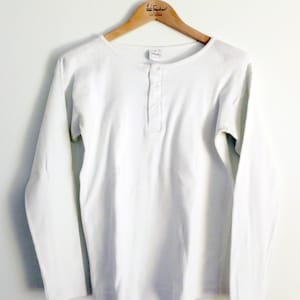 Könnte beinhalten: Ein weißes Langarm-Henley-Shirt mit gerippter Textur, das an einem Holzbügel hängt. Das Shirt hat eine Knopfleiste und einen Rundhalsausschnitt. Das Kleidungsstück besteht aus einem weichen, bequemen Material.