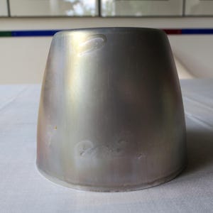 May include: A silver-colored, metallic, conical object with a wide base and a slightly flared top. The surface has subtle variations in color, with hints of gold and other hues. The object is likely a decorative piece or a functional item.