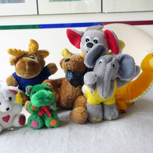 May include: A collection of plush toys including a brown moose, a green frog, a white teddy bear, a brown teddy bear, a gray elephant wearing a yellow tank top, a gray mouse with red ears, and a yellow dinosaur.