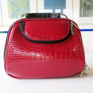 Puede incluir: Un bolso de mano rojo de piel sintética con textura de piel de cocodrilo. El bolso tiene un asa y ribetes negros, un pequeño bolsillo delantero y una cremallera plateada. El bolso está sobre una superficie blanca.