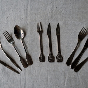 May include: A set of silverware, including forks, spoons, and knives, arranged in a semi-circle on a white tablecloth.
