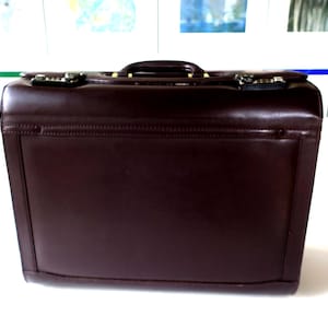 May include: A dark brown leather briefcase with a handle and metal clasps. The briefcase is rectangular with stitched edges. The handle is a darker brown than the main body. The briefcase is suitable for carrying documents.