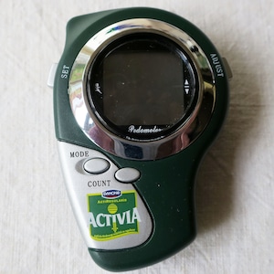 May include: A dark green pedometer with a silver bezel and a black display screen. The pedometer has buttons labeled "SET", "ADJUST", "MODE", and "COUNT". A sticker for "Activia" is visible on the device.