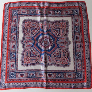 Può includere: Foulard quadrato in seta con un motivo paisley dettagliato in rosso, blu e bianco. La sciarpa ha un bordo rosso e un design centrale con intricate forme floreali e a goccia. Il design generale è simmetrico e ornato.