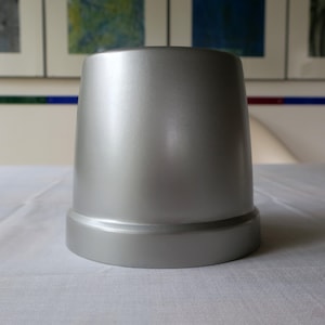 May include: A silver-colored, cylindrical object with a flared base. The object has a smooth, metallic finish and is set against a white tablecloth. The background features framed artwork.