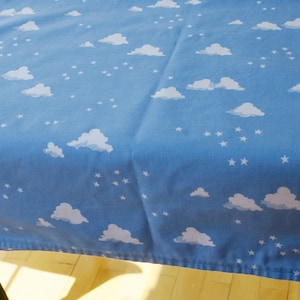 Könnte beinhalten: Eine blaue Tischdecke mit einem Muster aus weißen Wolken und Sternen. Der Stoff ist über einen Tisch drapiert und erzeugt eine skurrile Ästhetik. Das Design erinnert an einen klaren, sternenklaren Nachthimmel.