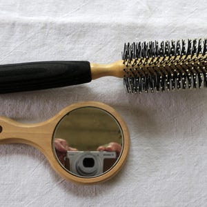 以下が含まれることがあります： 黒い木製の柄と黒い毛の丸いヘアブラシと、小さな丸い木製のハンドミラー。ブラシは毛のベースが明るい色です。鏡はカメラを映しています。