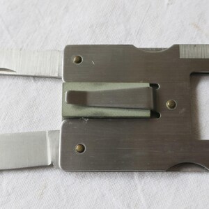 Puede incluir: Una tarjeta multiherramienta plateada con cuchillo, tenedor y abrebotellas. La tarjeta es rectangular con una ranura y un pequeño agujero. El cuchillo y el tenedor están desplegados. La herramienta es de metal con acabado cepillado.