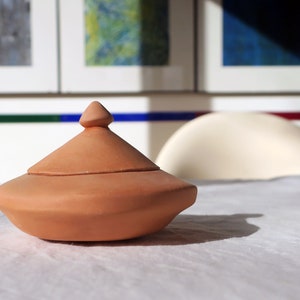 Könnte beinhalten: Eine tonfarbene, keramische Schale mit Deckel, abgerundetem, leicht ausgestelltem Boden und einer spitzen Oberseite. Die Schale steht auf einer weißen Oberfläche.