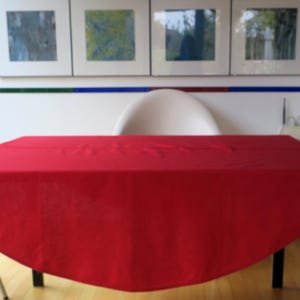 Könnte beinhalten: Ein rechteckiger Tisch mit einer einfarbigen roten Tischdecke. Der Tisch hat weiße Beine und einen schwarzen Trägerbalken. Der Hintergrund umfasst gerahmte Kunstwerke und einen weißen Stuhl.